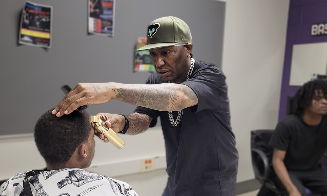A person sits in a chair wearing a plastic cape while a man wearing a cap holds clippers in front of a the person’s forehead.