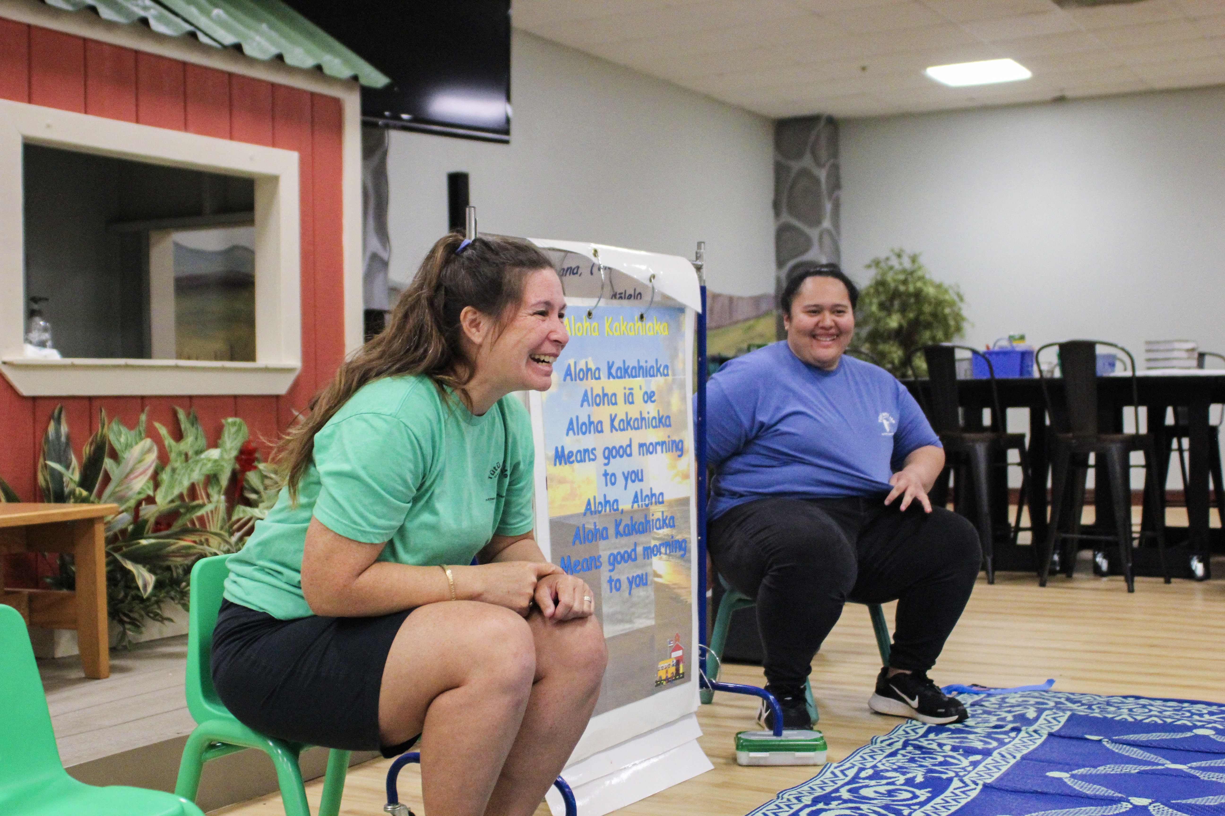 Two adults smile widely while sitting next to a sign with greetings written on it.