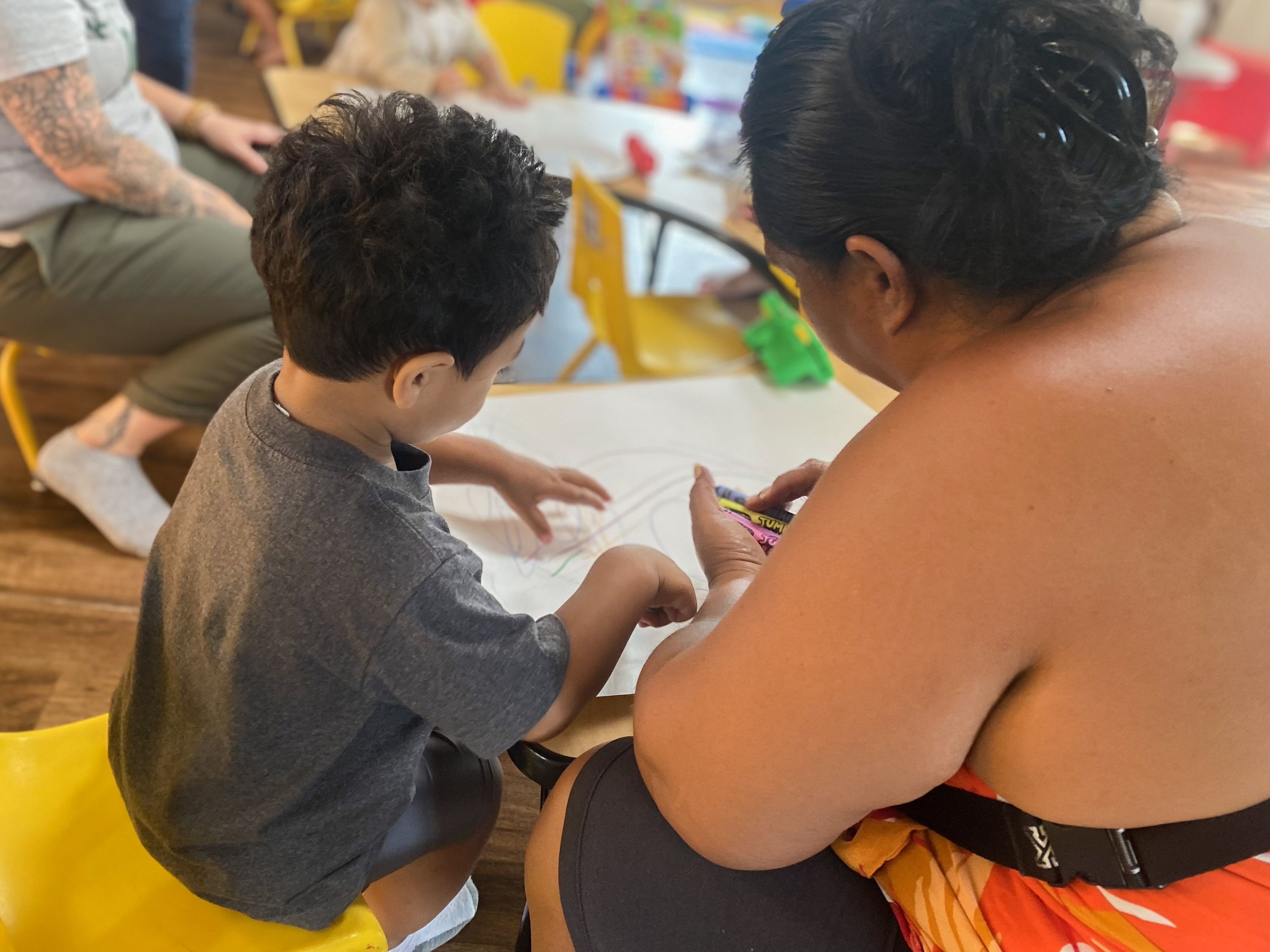 A woman sits with a child while they draw on paper.