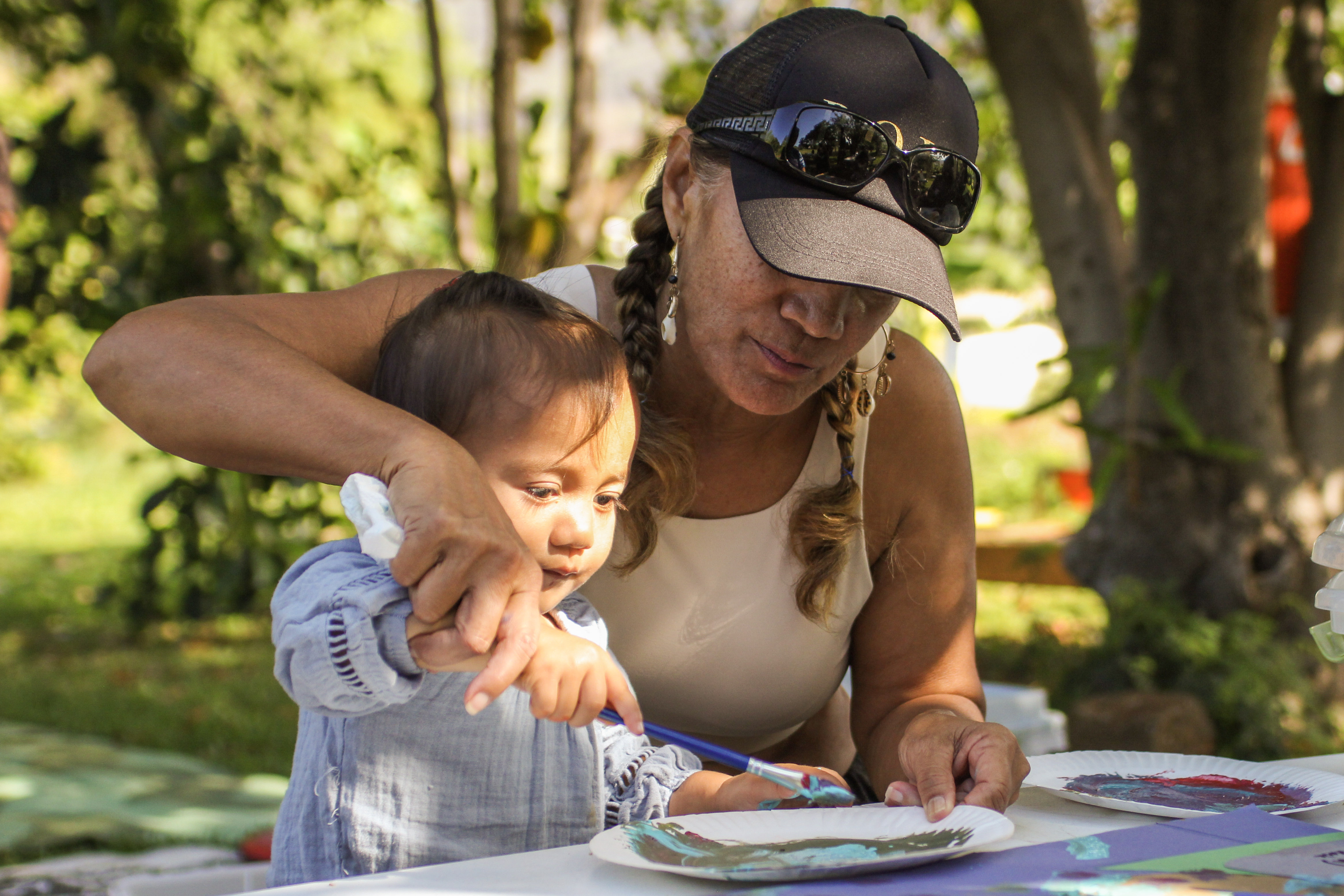 A person wearing a hat guides a child’s hand holding a paintbrush over a piece of paper on a table.