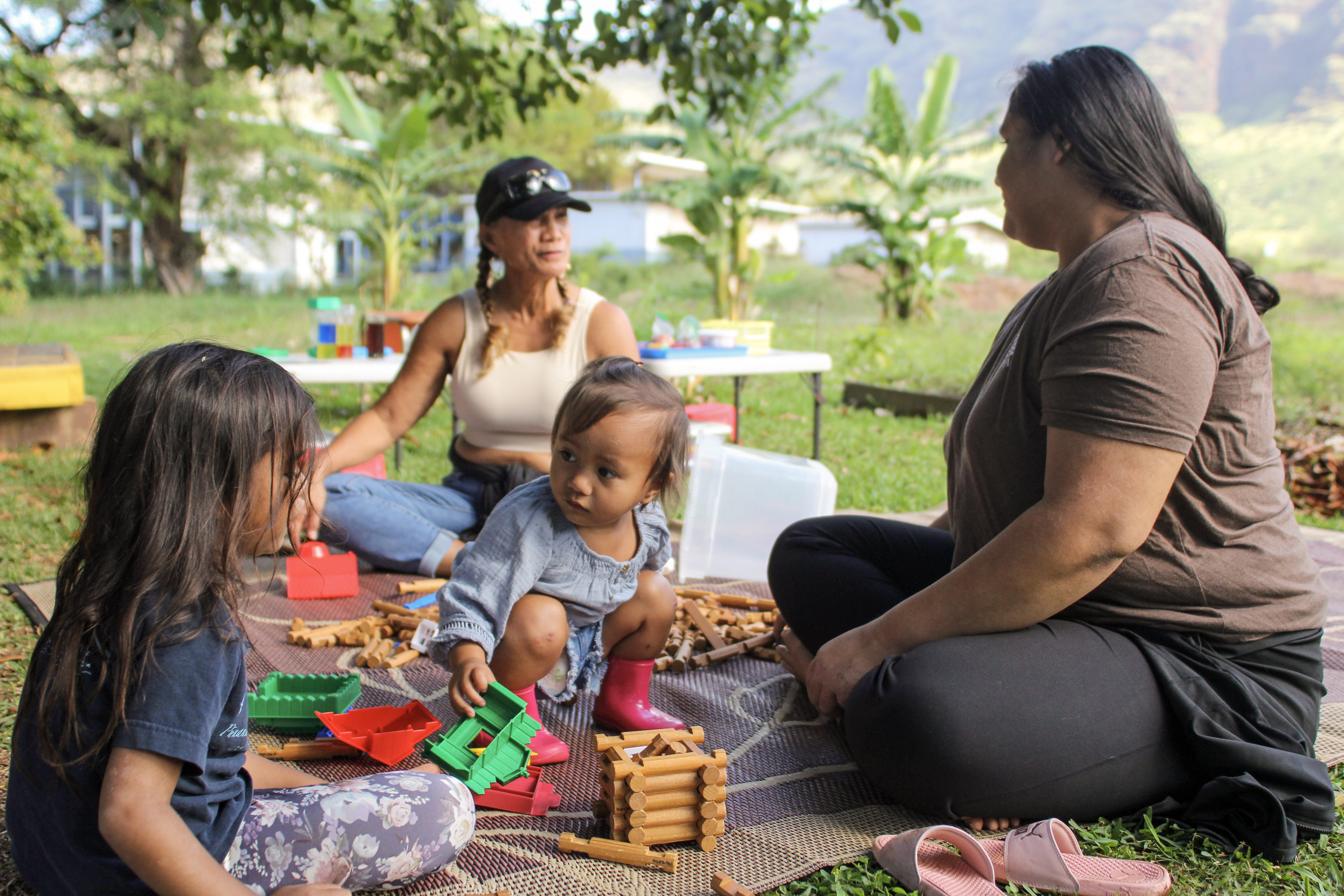 An adult sits with a child on a blanket spread on the grass outside, with a view of a hillside.