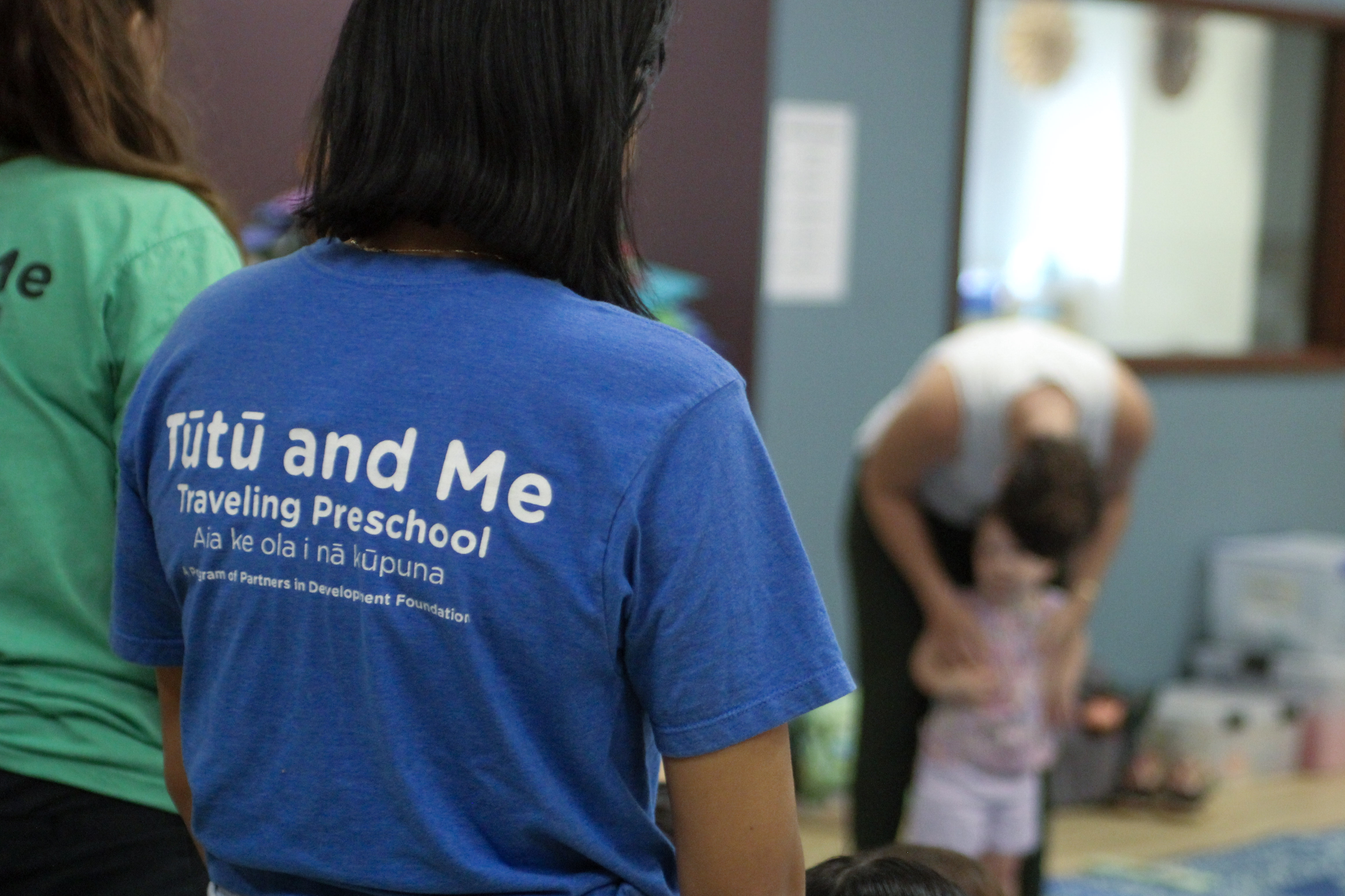 A person with a Tūtū and Me T-shirt looks at an adult interacting with a child.