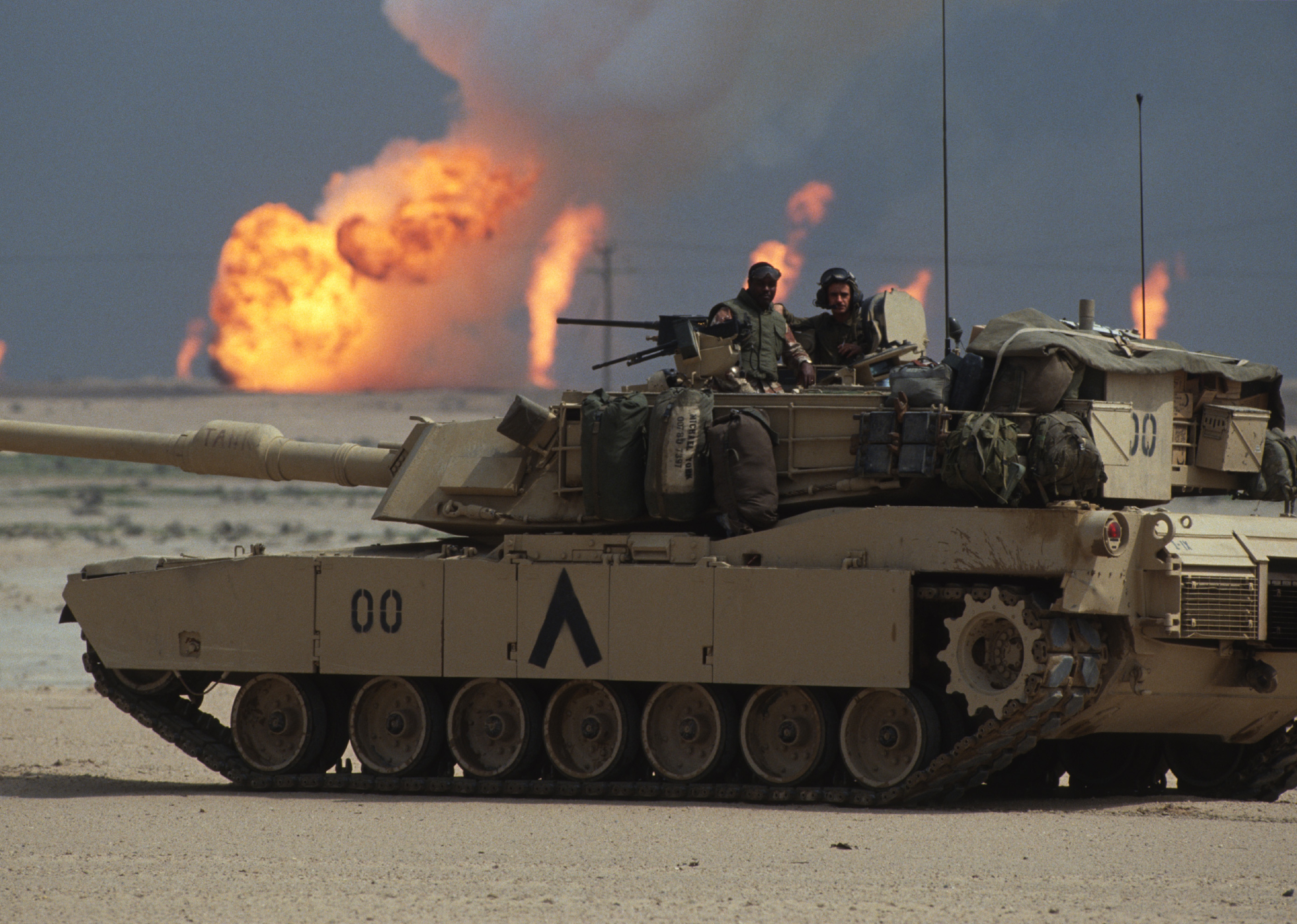 American soldiers stand in the turret of a tank as oil wells burn in the distance.
