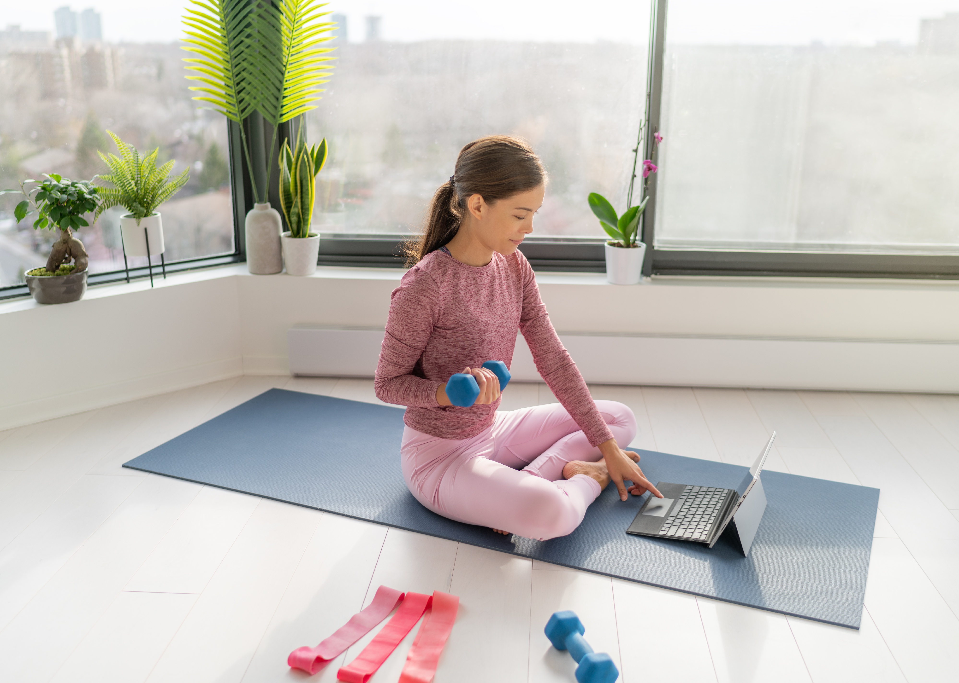 Woman at home sitting on yoga mat with a laptop in front of her, holding a dumbbell with another dumbbell and resistance bands beside her.