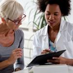 An older patient sits beside a doctor who is reviewing information on a clip board.