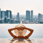 Woman wearing hat in urban infinity pool, looking out toward city buildings.