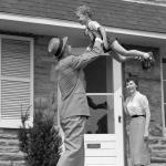 A 1950s father coming home and lifting his son in the air while his wife looks on from the doorway.