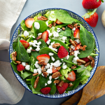 Strawberry, goat cheese, and walnut salad in a bowl, sits next to wooden spoon and dressing.