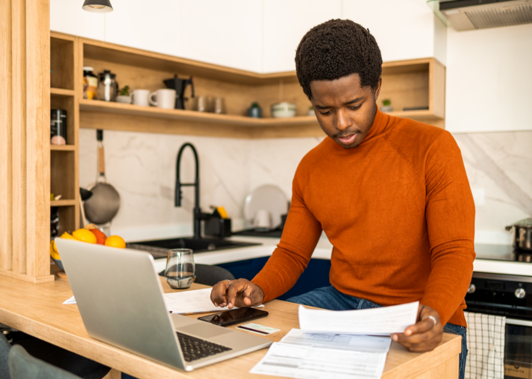 A man works on a laptop while looking at bills.