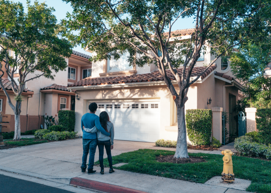 A couple wraps their arms around each other as they look at a house surrounded by trees.