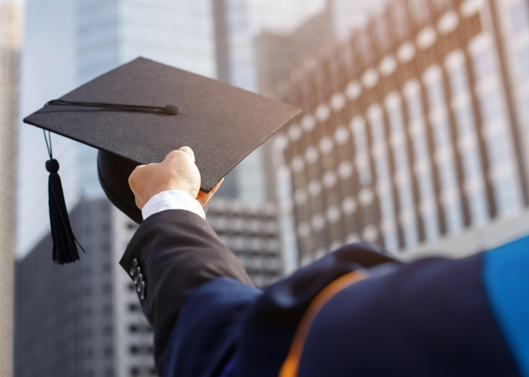 A college graduate holds their mortarboard aloft against a backdrop of office buildings.