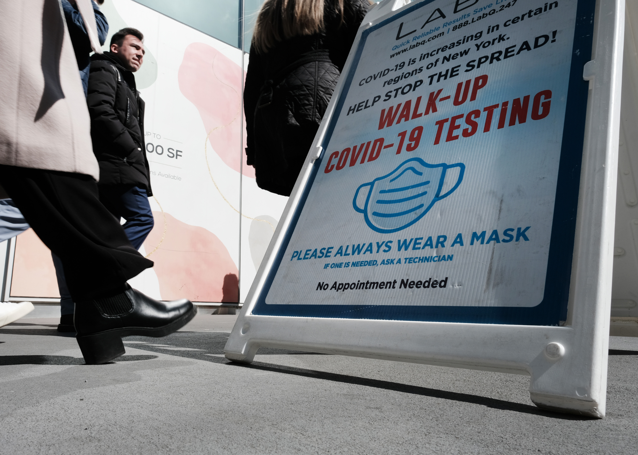 Photo shows a close up of a COVID-19 test site sign standing on a public sidewalk with people walking by. 