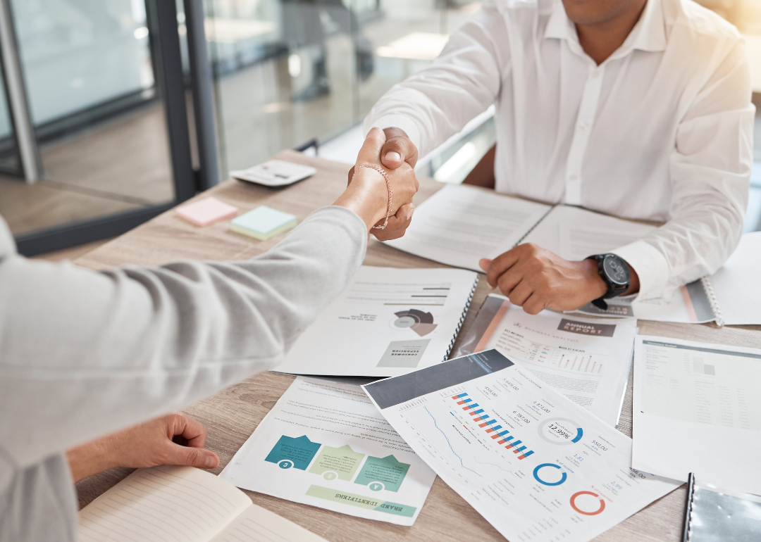 Two people shaking hands over an office desk with charts and reports.