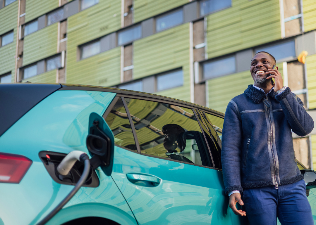 A man leans against an electric car talking on the phone as it charges. 