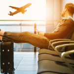 A man sits in a chair at an airport with his feet up on his carryon suitcase, watching a plane take off in the distance