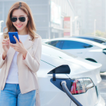 A woman looks at her smartphone while her white electric car charges at a public charge port.