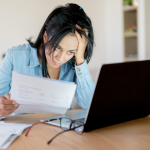A woman seated at a desk in front of a laptop, holing paperwork and looking frustrated.