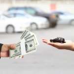 A conceptual image showing a financial transaction: one hand holding a small model car and another hand holding a stack of $100 dollar bills