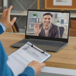 A office worker conducts a video job interview with a man shown on a laptop screen, while holding a copy of the man's resume