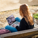 A young woman sits outside on a bench working on a laptop
