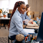 Two women sitting in a creative-looking office with plants and boxes, working on the computer and wrapping something in brown paper.