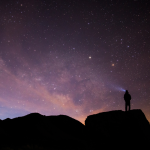 Silhouetted person stands atop boulder with light beam looking up at the milky way and sunrise.
