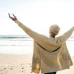 An older woman in a cardigan sweater stands on a beach with her arms raised