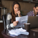 A young couple sit at a table. On the table is a laptop. The woman is holding a bill in her hands and looks worried. The man has has head in his hands.