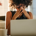 A Black woman sits in front of a laptop in a stressed posture, her hands covering part of her face