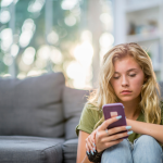 A young blonde girl sits in front of a gray couch and looks at her smartphone