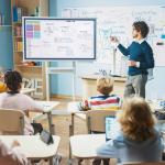 classroom from POV of students watching teacher at a screen in front