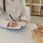 A doctor writes on a clipboard as they speak with a patient at a desk.