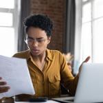 young person looks irritated while looking over paperwork in front of laptop with windows in background