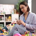 Young teacher sits in kindergarten classroom setting using smart phone