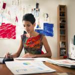 Business owner reviewing costs and paperwork sitting at desk with sewing machine and drawings visible behind her.