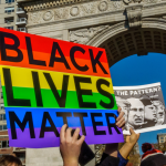Student protesters from NYU holding up a sign that says "Black Live Matter" with a Pride flag in the background, in Washington Square Park, New York City