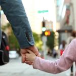 Adult holding child's hand waiting to cross a city street.