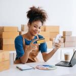 Woman holds credit card with one hand while reviewing purchases in small business setting with packages behind her. 