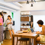 Three roommates share the kitchen, two cooking and one using a laptop at the table.