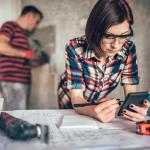 Two people in charge of overseeing a home improvement where one woman is leaning on a table with blueprints and noting with an instrument and the other is a man hammering nails on a wall.