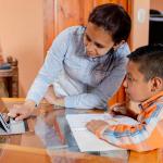 Adult helping child navigate a tablet set up at a table doing schoolwork in room with orange walls.
