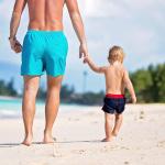 A dad and holding his toddler son's hand while they are walking on a white sand beach.