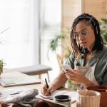 A woman with a gardening business is looking into a phone while writing down information on a notebook.