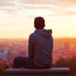 A man is sitting on a bench overlooking the city during sunset.