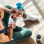 A young woman sitting on the floor post-workout and eating a bowl of oatmeal with fruits.