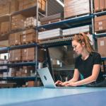 A woman managing logistics and using a laptop in a warehouse.