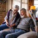An elder couple where the wife shows her phone to her husband who is holding on to a pair of crutches while sitting on a sofa.