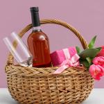 A Mother's Day gift basket with a wine bottle, wine glass, a gift box and pink flowers.
