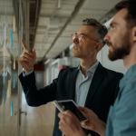 An older man in a suit and glasses standing and pointing to the glass in front of him while the other man is holding a digital screen looking up to the glass.