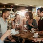 A group of six friends gathered around a wooden table in a cozy café. They are all smiling and laughing. Each person is holding a cup of coffee.
